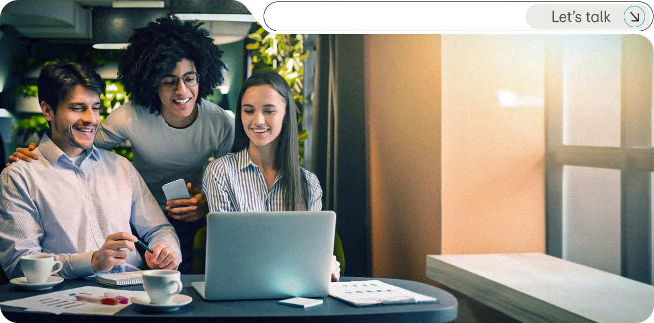 A diverse group of three professionals collaborating in a modern workspace, smiling and engaged around a laptop.